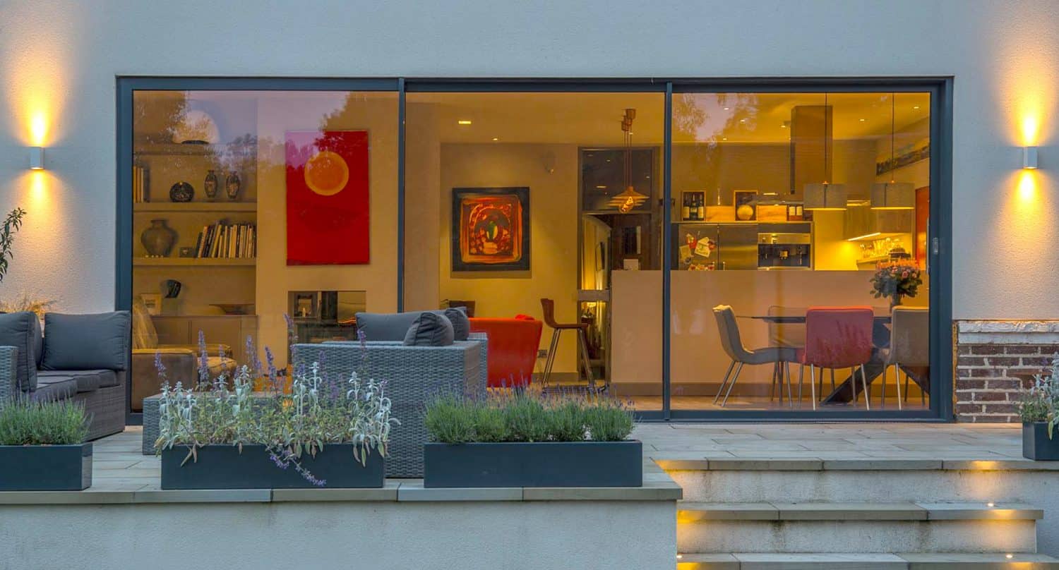 Modern home with large glass sliding doors revealing a warmly lit interior, including a kitchen and dining area. Outdoor patio features gray seating, potted plants, and steps leading up to the house.
