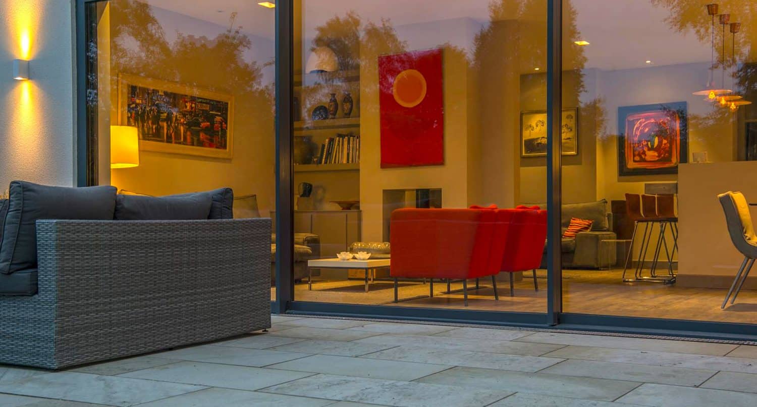 Modern living room viewed through large glass doors, featuring red chairs, wall art, a gray sofa, and warm lighting. On the patio outside, there is a gray wicker sofa with dark cushions and stone tile flooring.