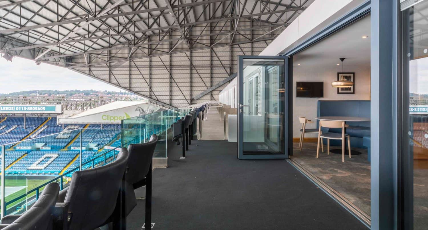 A modern stadium suite with glass doors open to a private balcony, offering cushioned seating and a clear view of the football field and stands below.