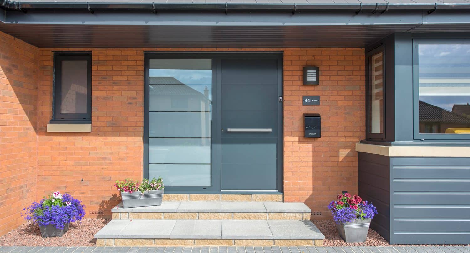Modern house entrance with a sleek gray door, frosted glass panels, brick walls, a mailbox, house number sign, and potted flowers on either side of the steps leading up to the door.