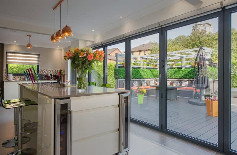 Modern kitchen with glass doors opening to a spacious patio. The kitchen features an island with flowers, barstools, and pendant lights, while the patio has seating, plants, and umbrellas, with houses and greenery in the background.