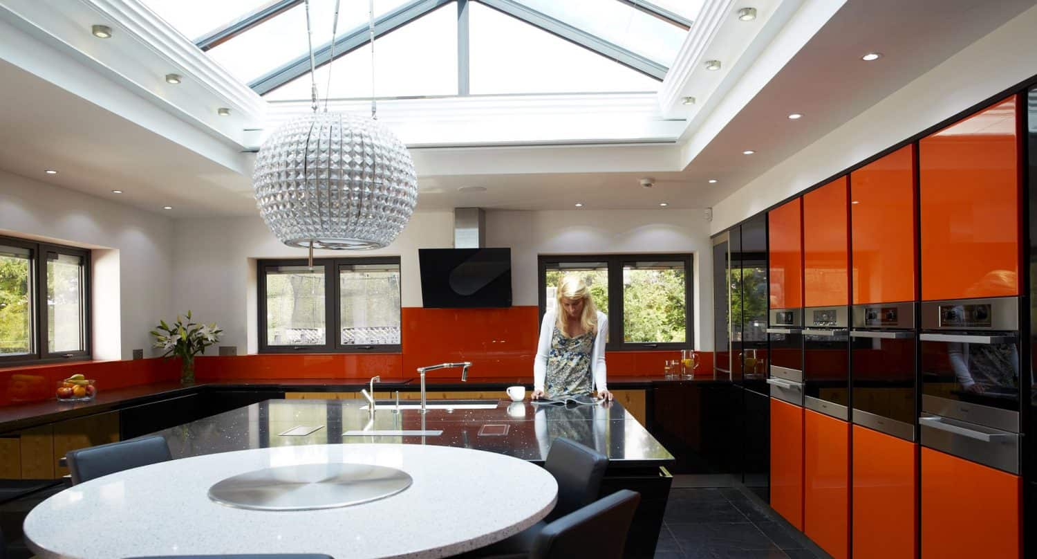 A modern kitchen with orange and black cabinets, a large skylight, a round white island with seating, and a woman standing near the sink. A large decorative pendant light hangs over the island.