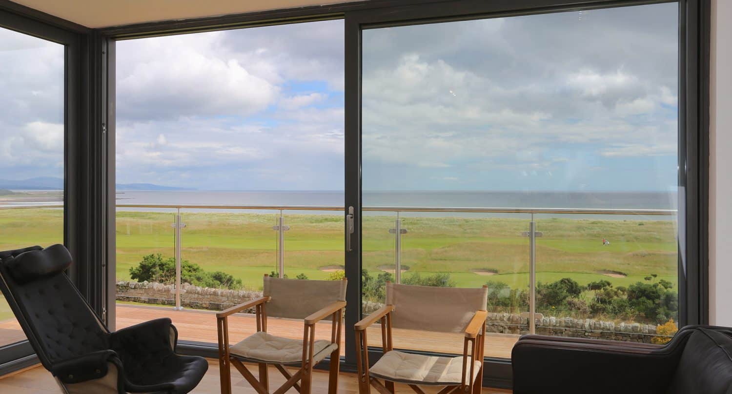 A bright room with large glass doors opens to a balcony, showing a scenic view of grassy fields, a sandy shoreline, and a calm sea under a partly cloudy sky. Two wooden chairs and a black chair face the view.