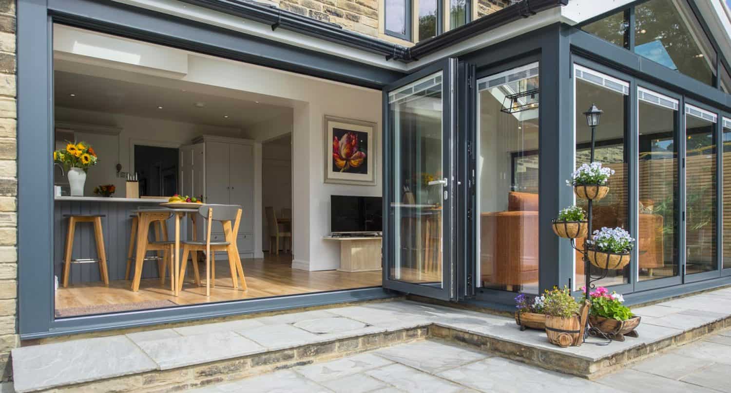 Modern home with large glass folding doors opening to a patio. Inside is a bright kitchen with wood chairs, sunflowers on the counter, and a painting on the wall. Outside, there are potted flowers on stands by the entrance.