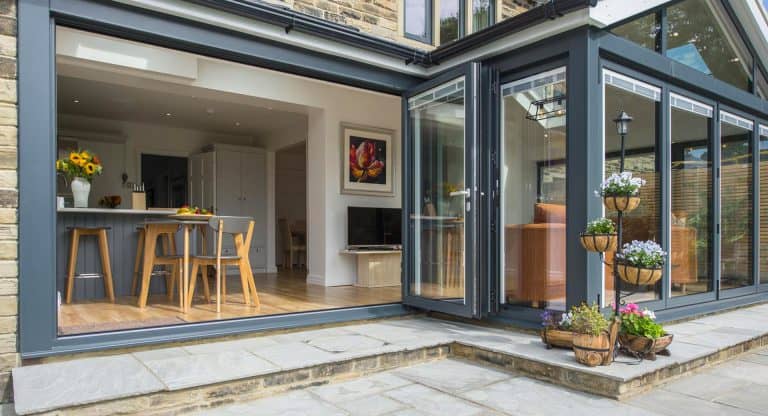 Modern home with large glass folding doors opening to a patio. Inside is a bright kitchen with wood chairs, sunflowers on the counter, and a painting on the wall. Outside, there are potted flowers on stands by the entrance.