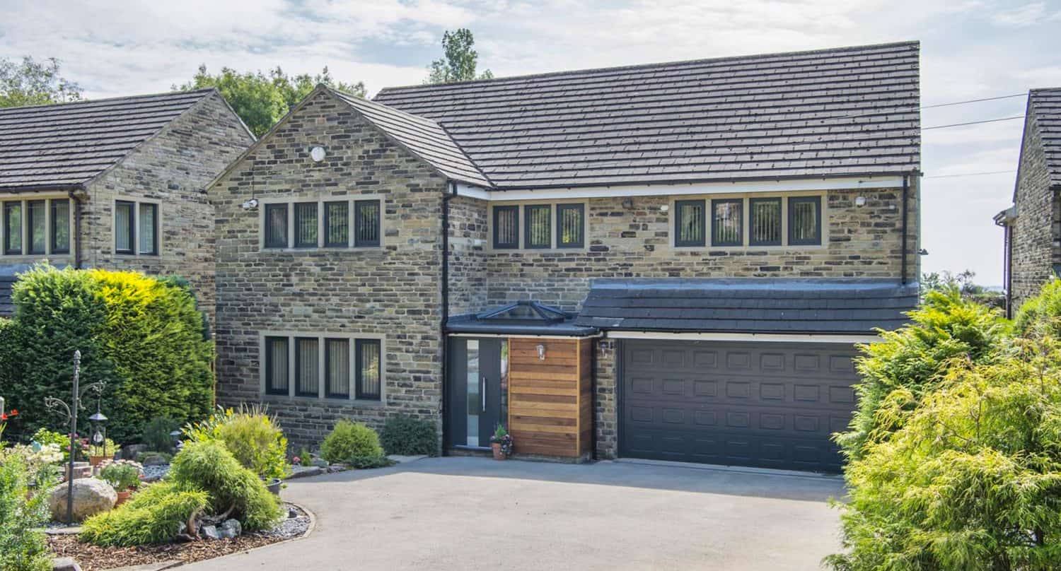 Modern two-story stone house with multiple windows, a black garage door, and a wooden accent. There is a small garden with shrubs and flowers in front, and a driveway leading to the garage.