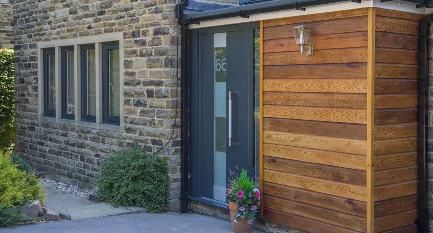 Modern house entrance with a glass front door displaying the number 66, surrounded by stone walls and wood paneling. A potted plant with flowers sits by the door, and greenery lines the wall.