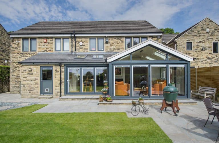 A modern two-story stone house with large glass windows and doors, a green lawn, patio furniture, potted plants, and a barbecue grill in the backyard under a partly cloudy sky.