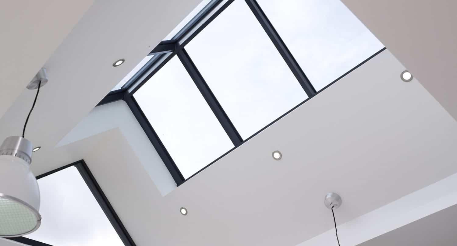 A modern ceiling with a large rectangular skylight and triangular window allows natural light into the minimalist white interior, complemented by recessed lights, hanging silver pendant lights, and sleek bifold doors.