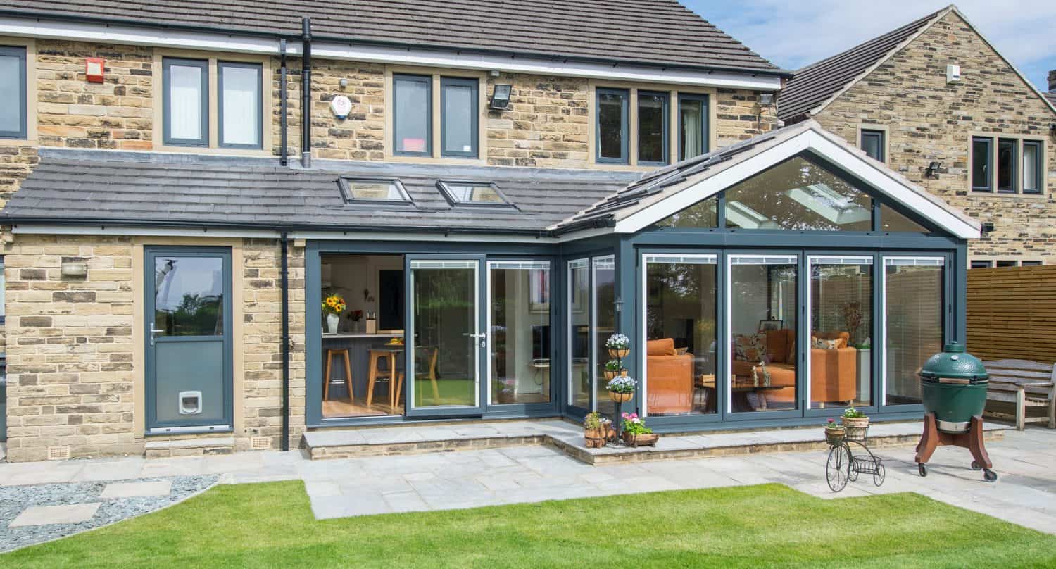 Modern stone house with a glass sunroom extension, featuring large windows and sliding doors opening onto a neatly trimmed lawn and patio with outdoor furniture and a barbecue grill.