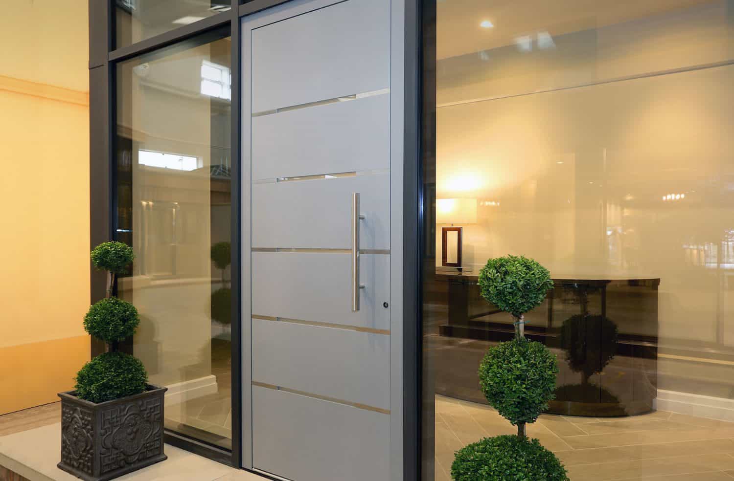 Modern gray aluminium front door with horizontal metal lines, flanked by large glass windows and two decorative potted topiary plants on either side, inside a well-lit contemporary entryway.