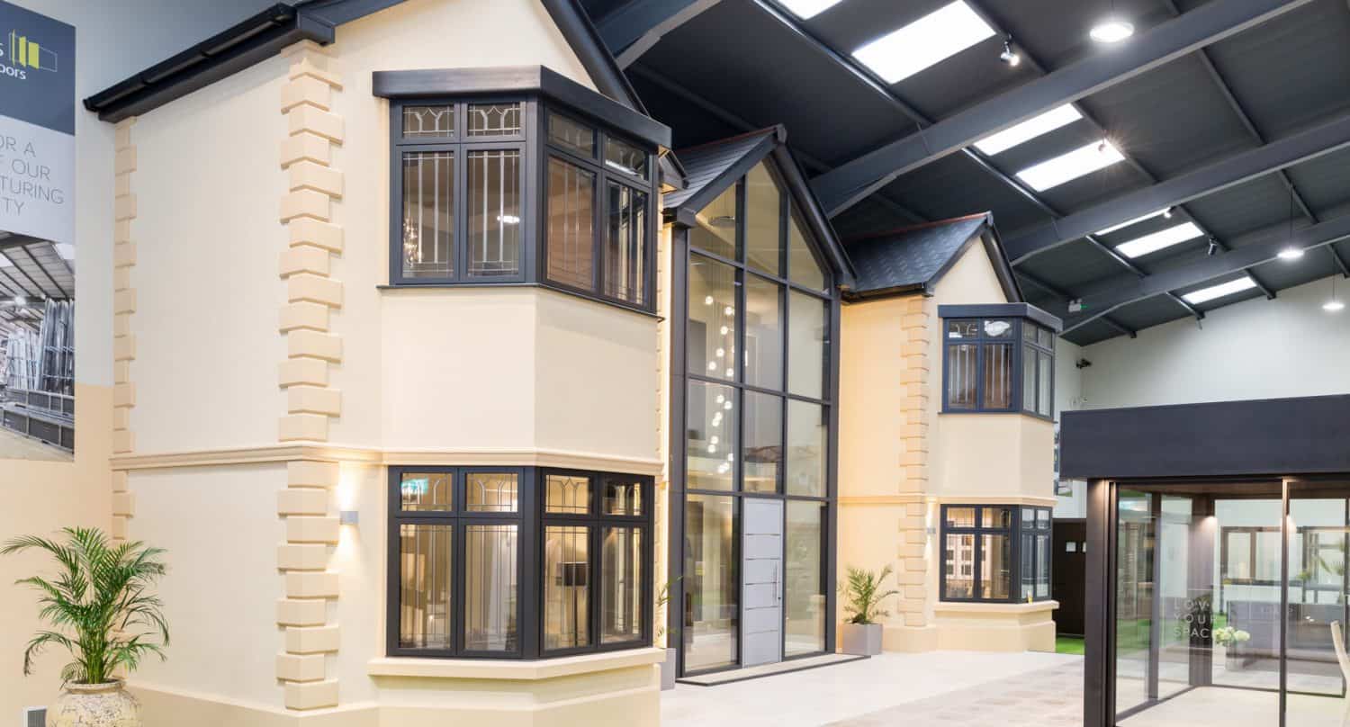 A modern showroom displays a full-scale model of a two-story cream house with black-framed bay windows, glass double doors, and large windows, all under a high industrial roof with skylights.