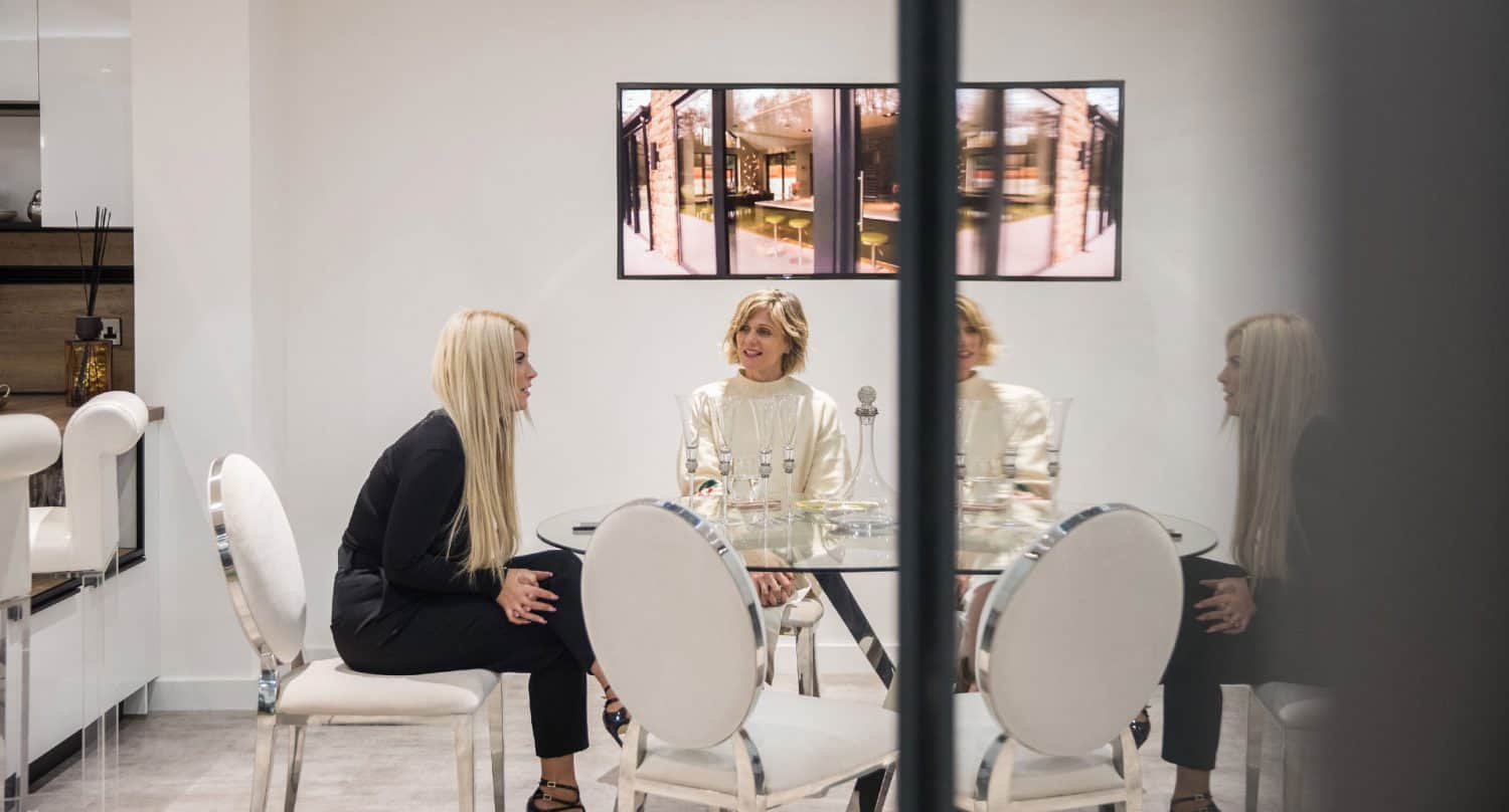 Two women sit and talk at a round glass table with white chairs in a modern, bright room featuring bespoke glass solutions. Artwork hangs on the wall behind them, and their reflections are visible in a mirror.