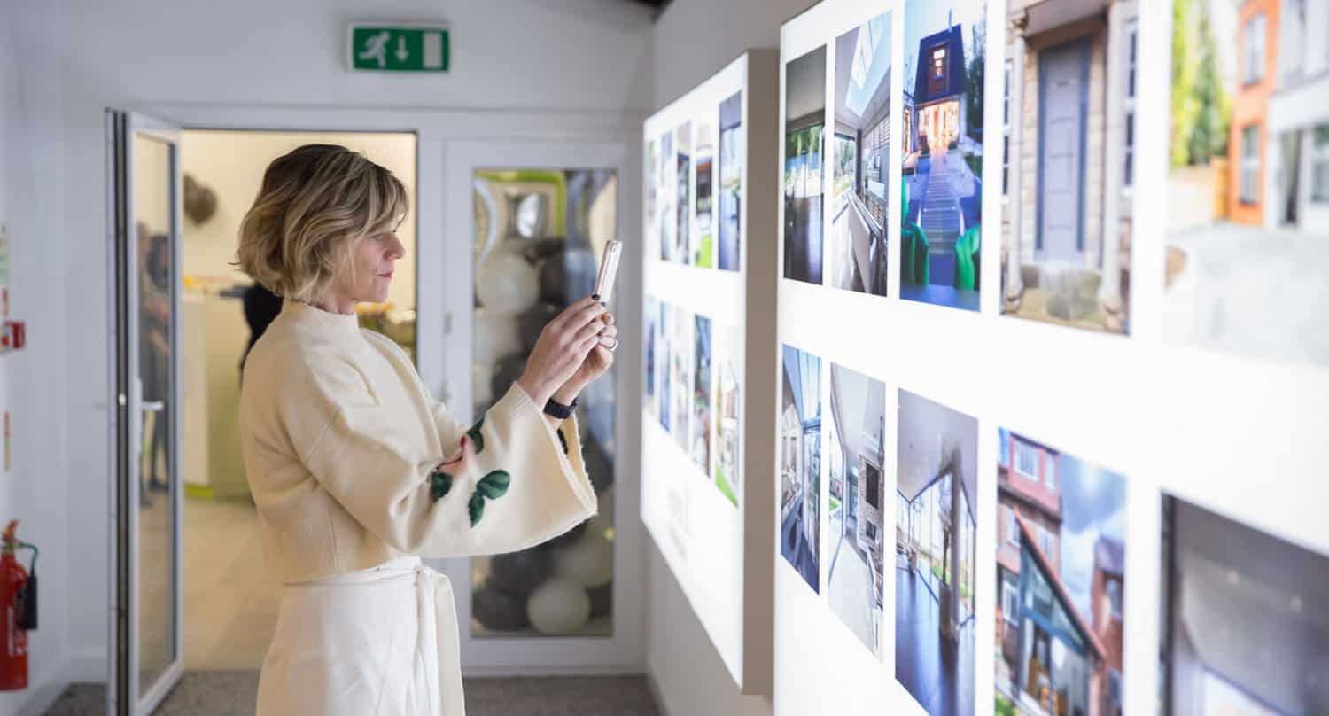 A woman in a light-colored outfit uses her smartphone to capture illuminated images displayed on a gallery wall beneath striking glass roofing.