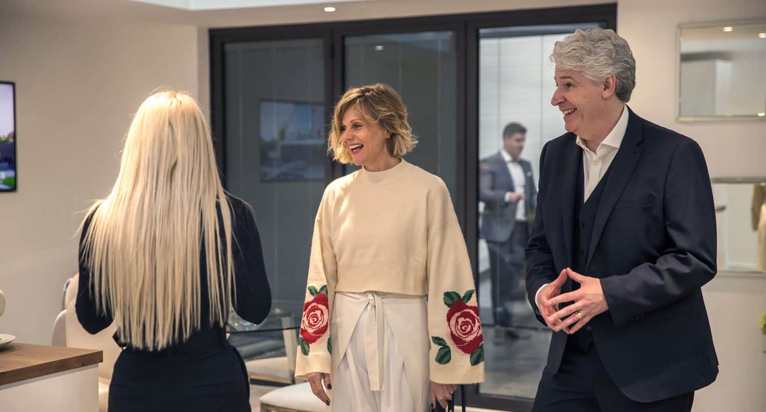 Three people are talking indoors near large windows featuring bespoke glass solutions. A woman with long blonde hair, seen from behind, faces a smiling woman in a white sweater with red roses and a man in a suit. Another person is visible outside through the window.