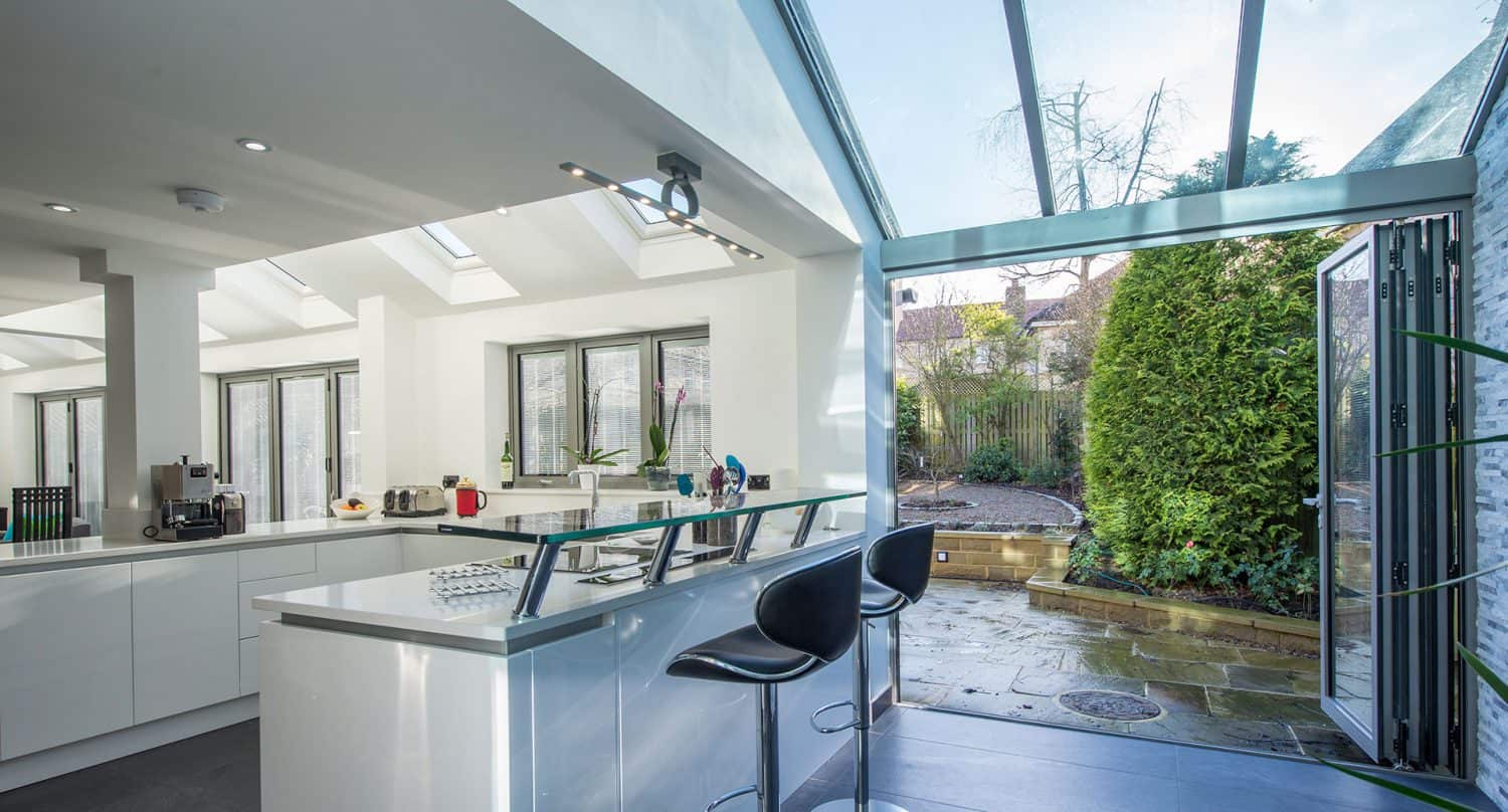 Modern kitchen with white cabinets and an island with two black bar stools, featuring bespoke glass solutions like large glass doors that open to a patio and lush green garden. Natural light fills the space through skylights and windows.