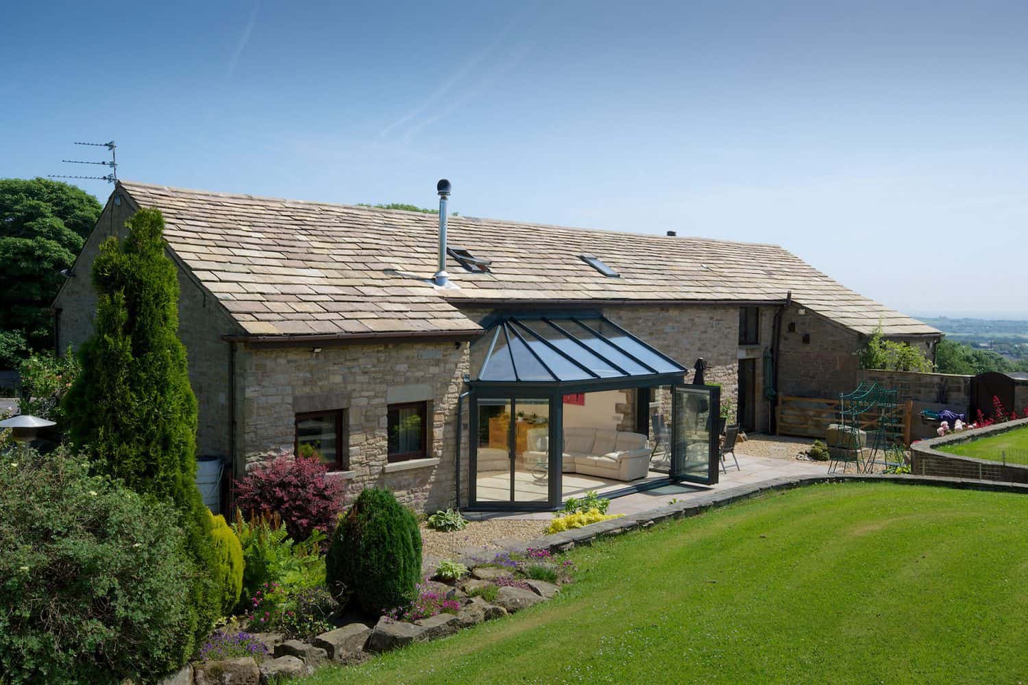A large stone house with a glass sunroom extension featuring bespoke glass solutions, surrounded by a well-kept lawn, shrubs, and colorful garden beds, set against a clear blue sky and distant countryside views.