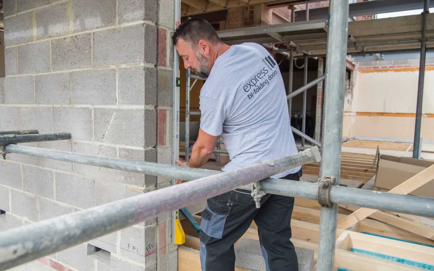 A construction worker in a gray express bi-fold doors t-shirt uses a tool on a partially built house, surrounded by scaffolding and unfinished brick walls.