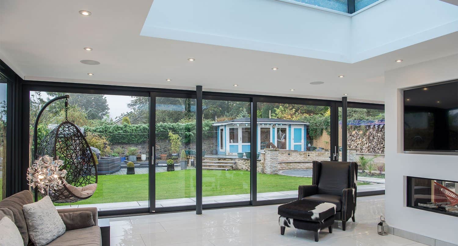Modern living room with large bifold doors overlooking a green garden and a blue gazebo. The room features a hanging chair, armchair, fireplace, and tiled floor under a skylight.