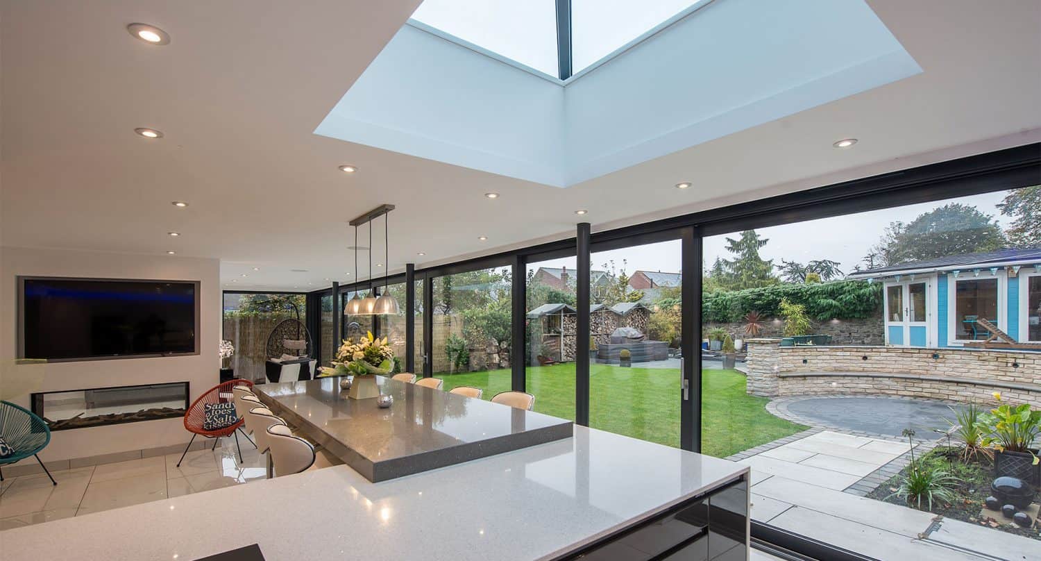 Modern kitchen with a large island, skylight, recessed lighting, and floor-to-ceiling bifold doors opening to a spacious, landscaped backyard with green lawn and a contemporary garden house.