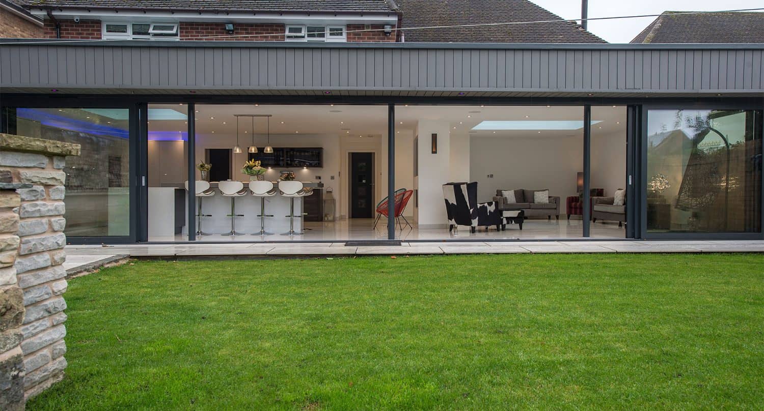 Modern house with bespoke glass solutions, featuring large sliding doors opening to a green lawn; inside, a white kitchen with bar stools, dining area, and stylish sitting area with black and white chairs are visible.