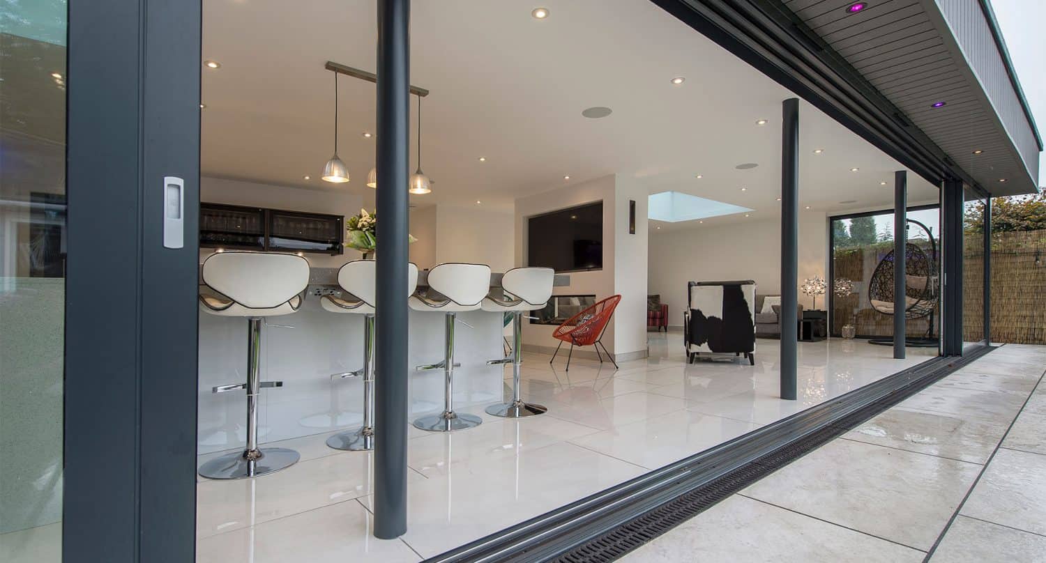 Modern open-plan kitchen and living space with white tile flooring, a row of bar stools at a counter, pendant lights, and large bifold doors opening onto a patio.