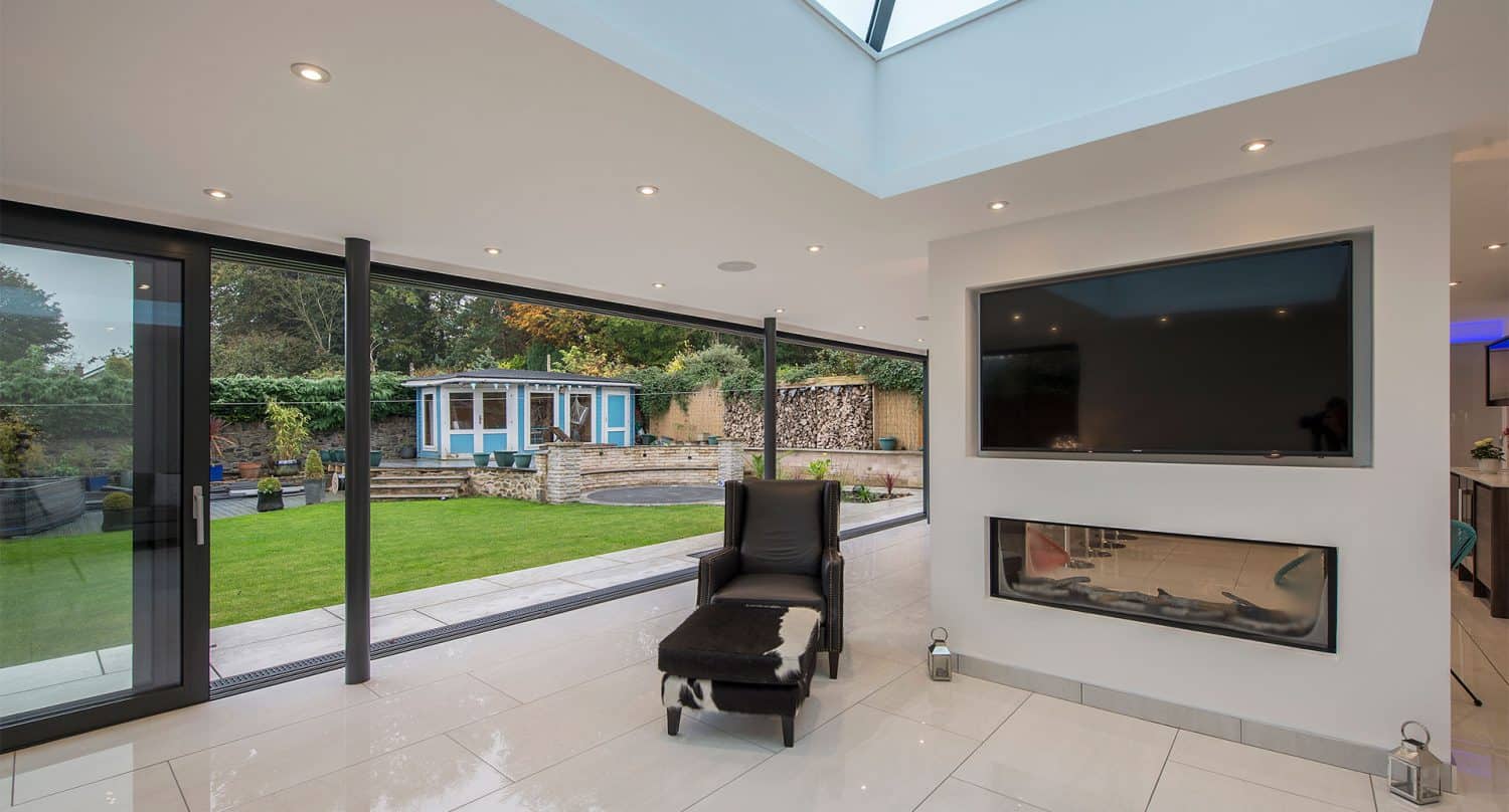A modern living room with large sliding doors and glass walls overlooking a green garden and a blue garden house. Inside, there’s a black chaise lounge, a cowhide ottoman, a wall-mounted TV, and a built-in fireplace.