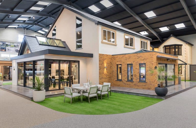 A modern house with large windows and a mix of brick and cream exterior is displayed indoors. Outdoor furniture and plants are arranged on artificial grass under a high, industrial-style roof.