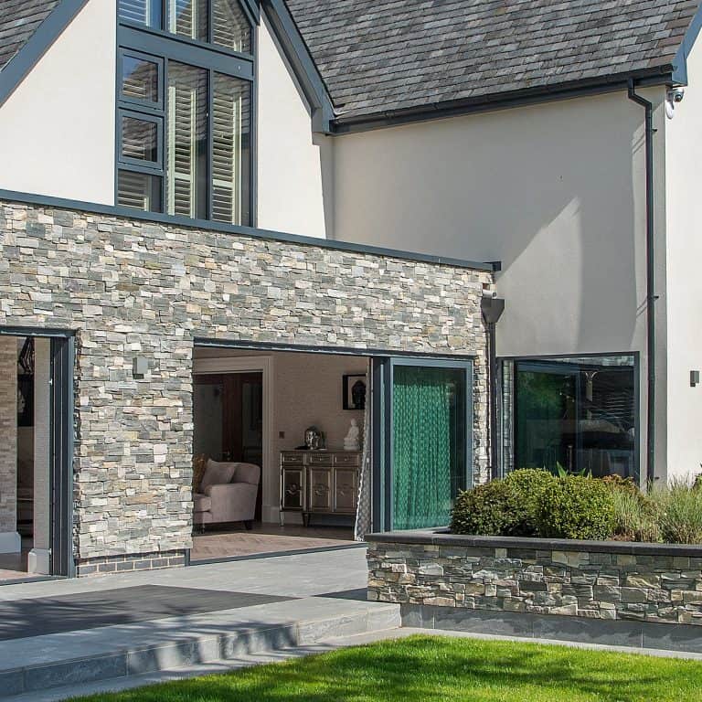 Modern house exterior with large windows, stone and cream walls, patio doors opening to a living area, and a landscaped garden with green grass and bushes in the foreground.