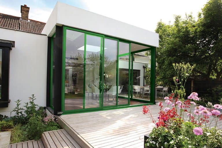 A modern house extension with floor-to-ceiling green-framed glass doors opens onto a wooden deck surrounded by colorful flowers and greenery, with trees and outdoor furniture visible in the background.