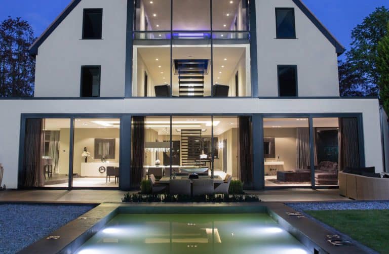 Modern three-story house with large glass windows and doors, brightly lit interior, visible staircase, outdoor seating area, pool in the foreground, and trees surrounding the property at dusk.