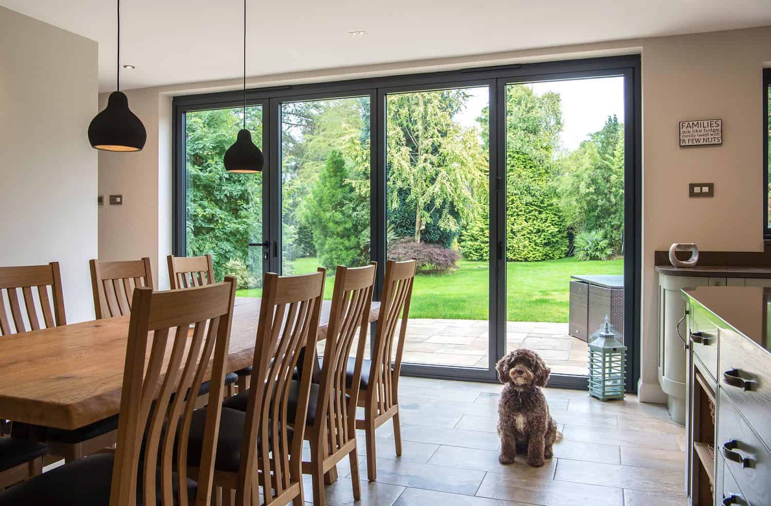 A cozy dining area with a wooden table and chairs, large glass doors overlooking a green garden, and a small brown dog sitting on the tiled floor near the windows.