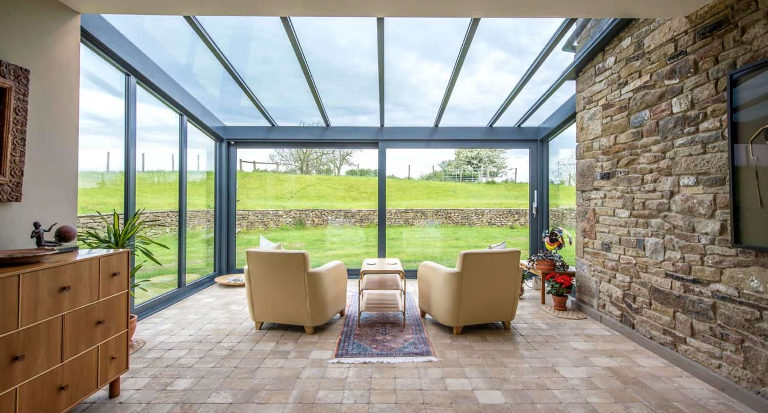 A bright sunroom with glass walls and ceiling, two beige armchairs, a small table, stone floor, potted plants, and a view of a green grassy field outside.