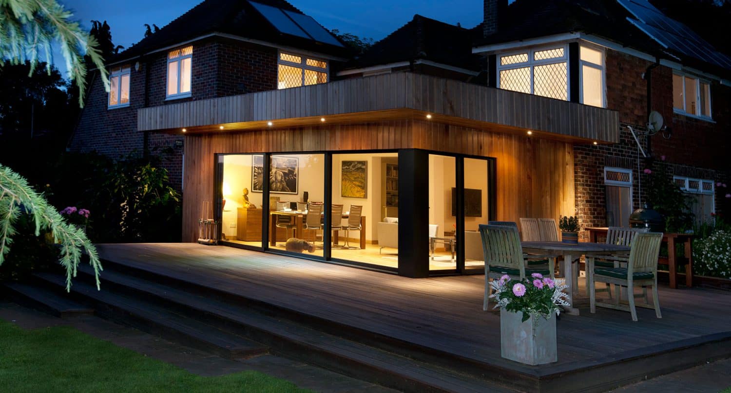 Modern wooden house extension with large glass sliding doors, warmly lit interior with furniture visible. Outdoor wooden deck has a dining table, chairs, and potted plants, evening sky in the background.