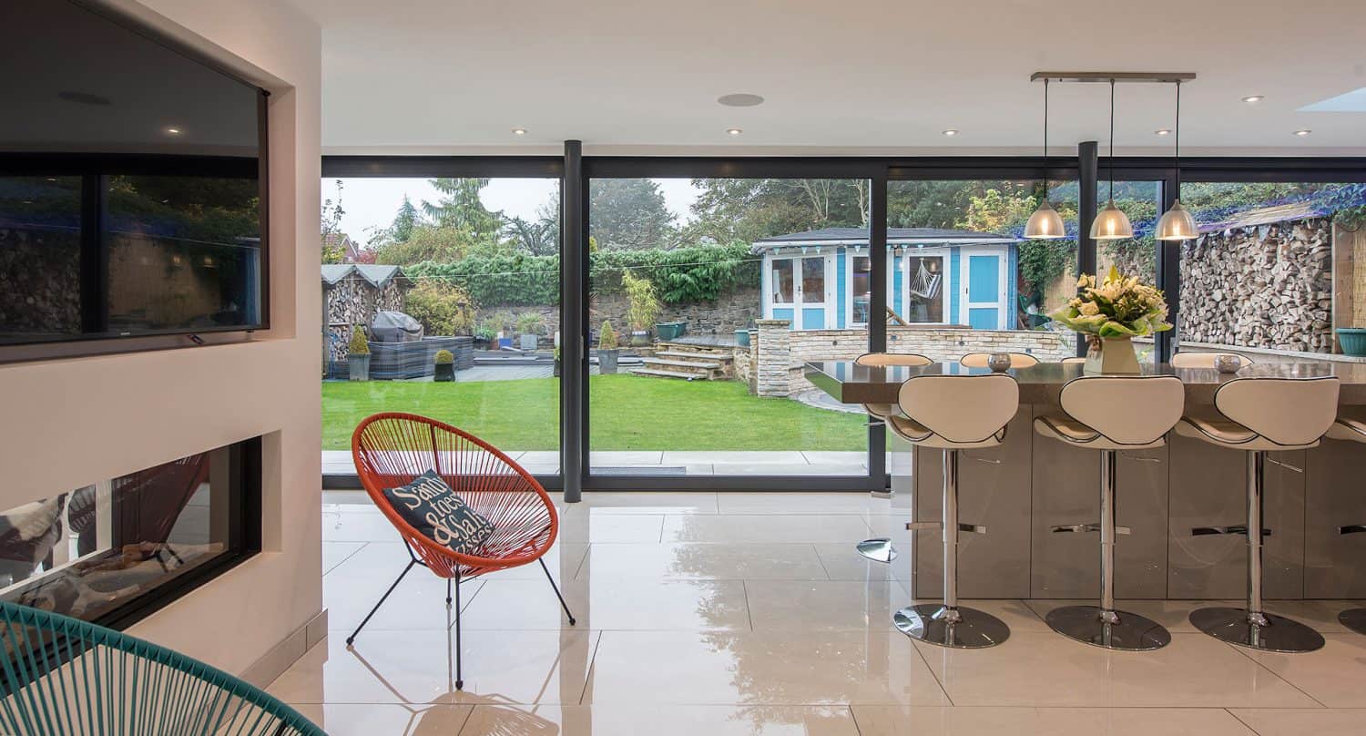 Modern kitchen with large bespoke glass sliding doors overlooking a landscaped garden. Features a breakfast bar with white stools, pendant lights, and a red accent chair. Outside, there is a blue garden shed and stacked firewood.