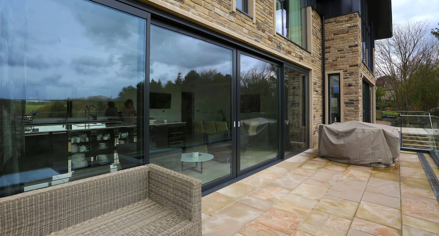 Modern patio with beige stone tiles, a covered barbeque grill, and a wicker chair. Large glass sliding doors reflect trees and sky, giving a glimpse into a contemporary kitchen and dining area inside.