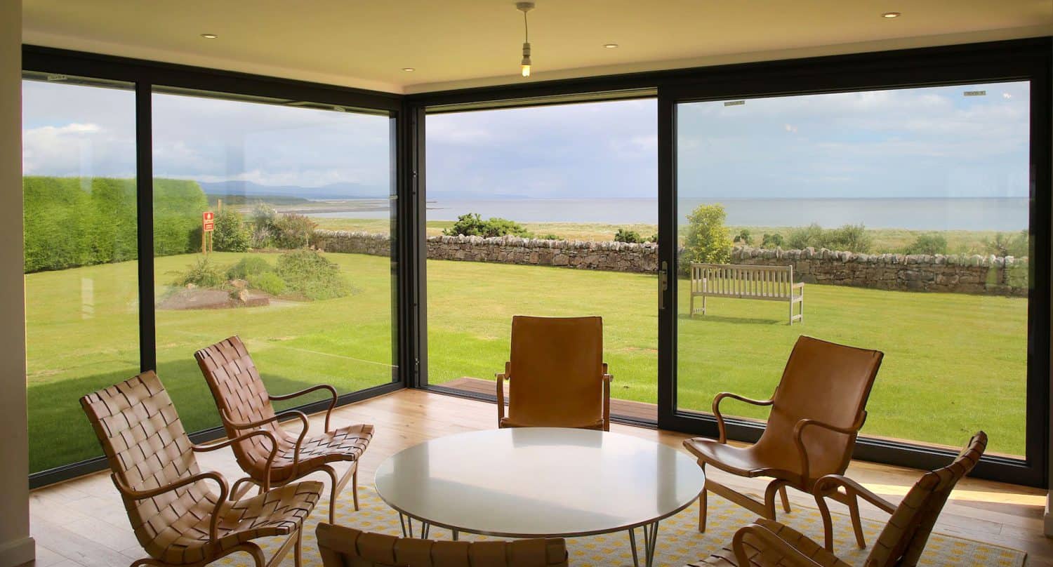 A modern sunroom with large floor-to-ceiling windows offers a scenic view of a green lawn, stone wall, and the sea beyond. The room has four tan leather chairs around a white oval table.
