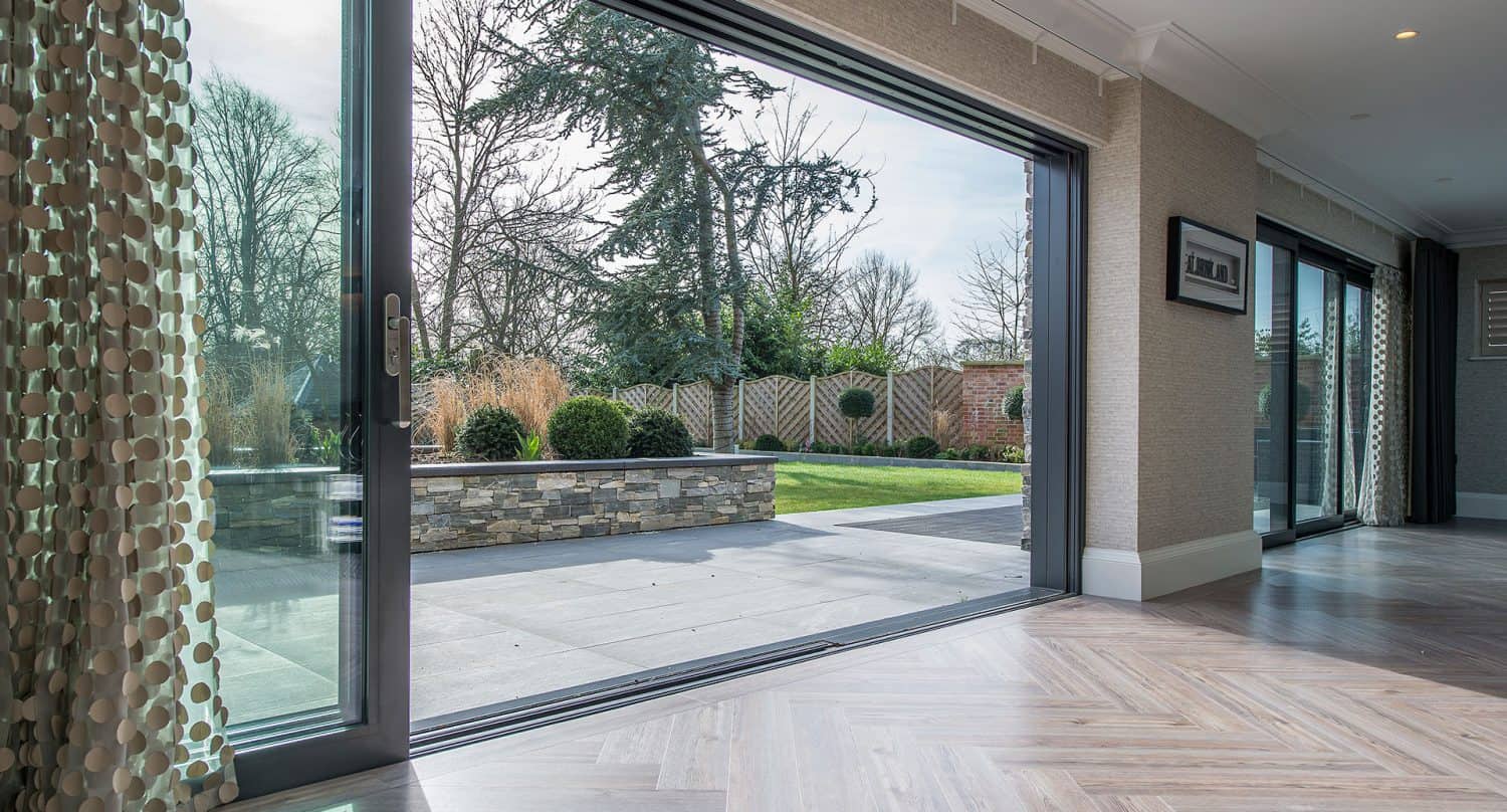 A large glass sliding door opens from a modern home interior with wooden floors to a spacious, landscaped backyard with a stone patio, trimmed bushes, trees, and a wooden fence.