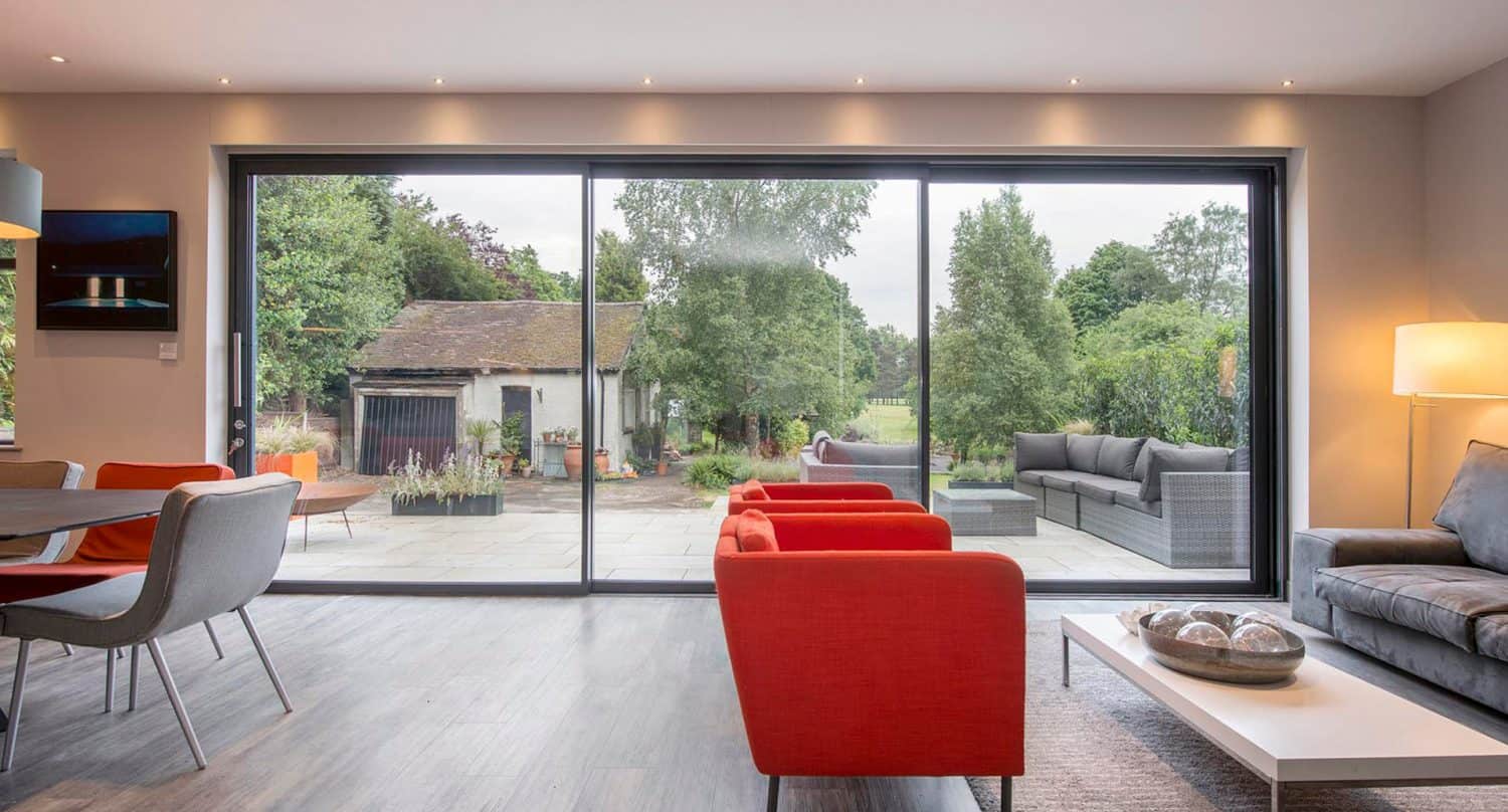 Modern living room with red and gray seating, large sliding glass doors opening to a patio with outdoor furniture, and a garden view with trees and a rustic shed.