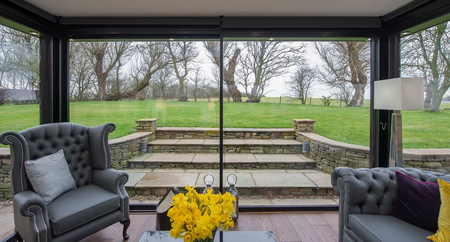 A modern sunroom with large glass windows, two gray armchairs, a side table with yellow flowers, and an outdoor view of stone steps leading to a green lawn and leafless trees.