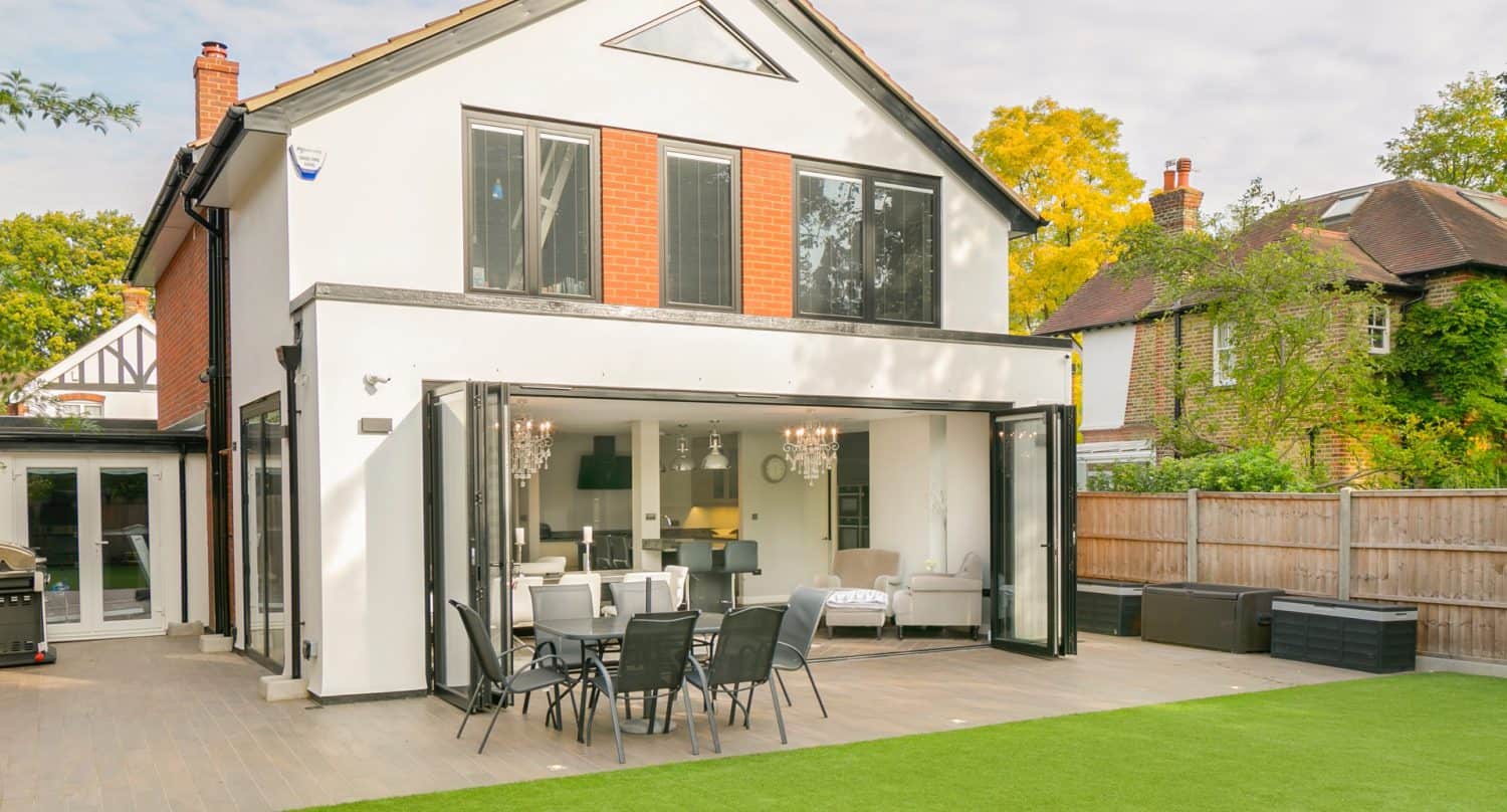 Modern two-story house with large glass folding doors open to a patio, outdoor dining set, and a lawn; interior has chandeliers, sofas, and a kitchen visible through the open space.