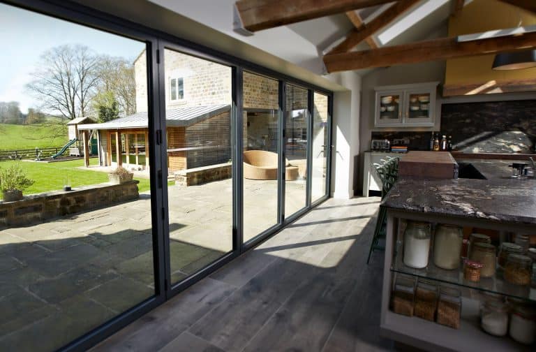 Modern kitchen with large glass sliding doors opening to a patio and grassy yard. Exposed wooden beams, marble countertop, and jars on shelves add a rustic touch. Outdoor seating and a wooden structure are visible.