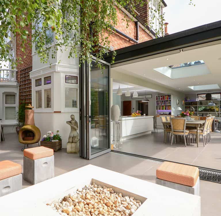 Modern open-plan kitchen and dining area with sliding glass doors leading to a patio. The patio features a fire pit, outdoor seating, sculptures, and lush greenery, seamlessly blending indoor and outdoor spaces.