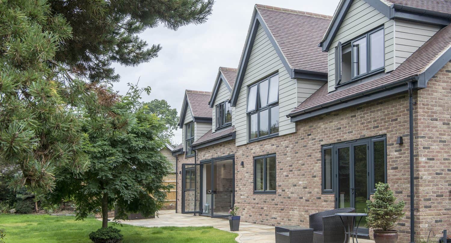 A modern brick house with large windows, sliding doors, and gable roofs, featuring a patio with outdoor furniture and potted plants, surrounded by a green lawn and trees.