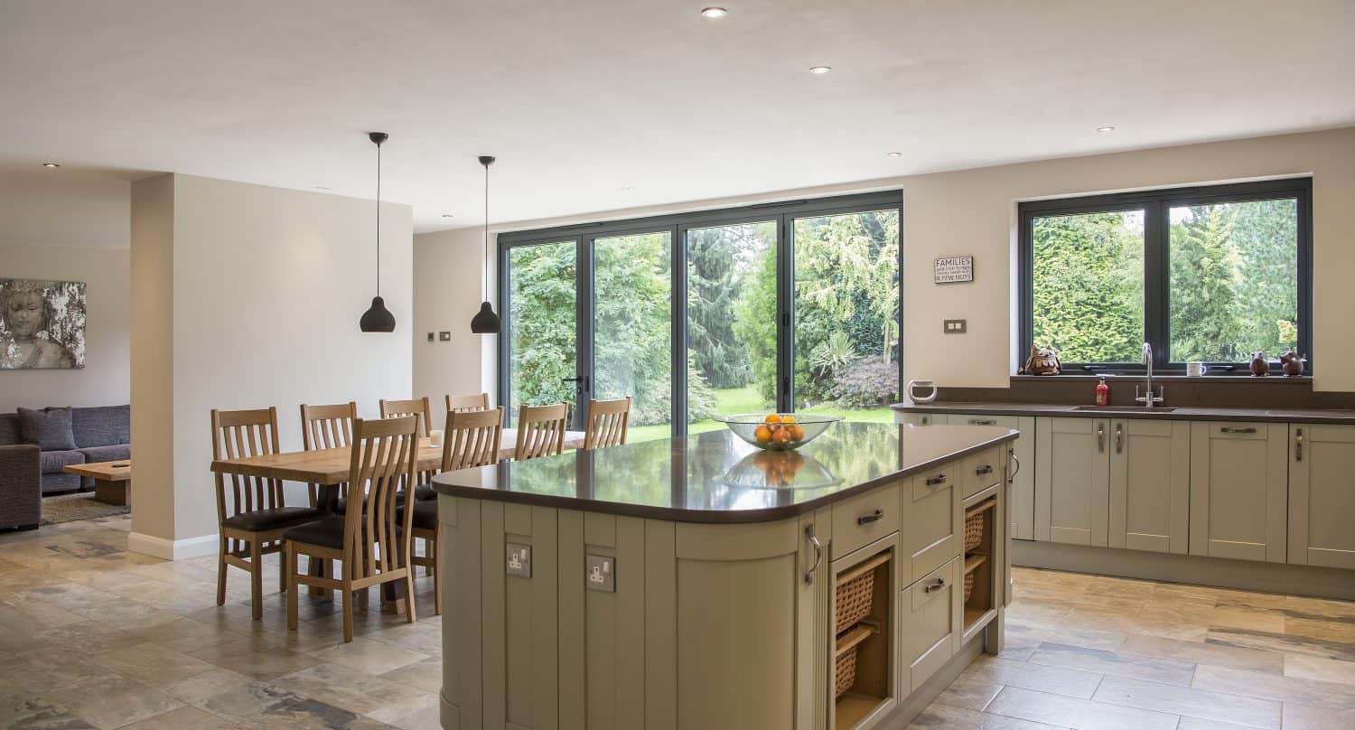 Spacious kitchen with a large island, wooden dining table for eight, pendant lights, and wide windows offering a garden view. Bifold doors seamlessly connect the modern, neutral-toned space to the lush outdoors.