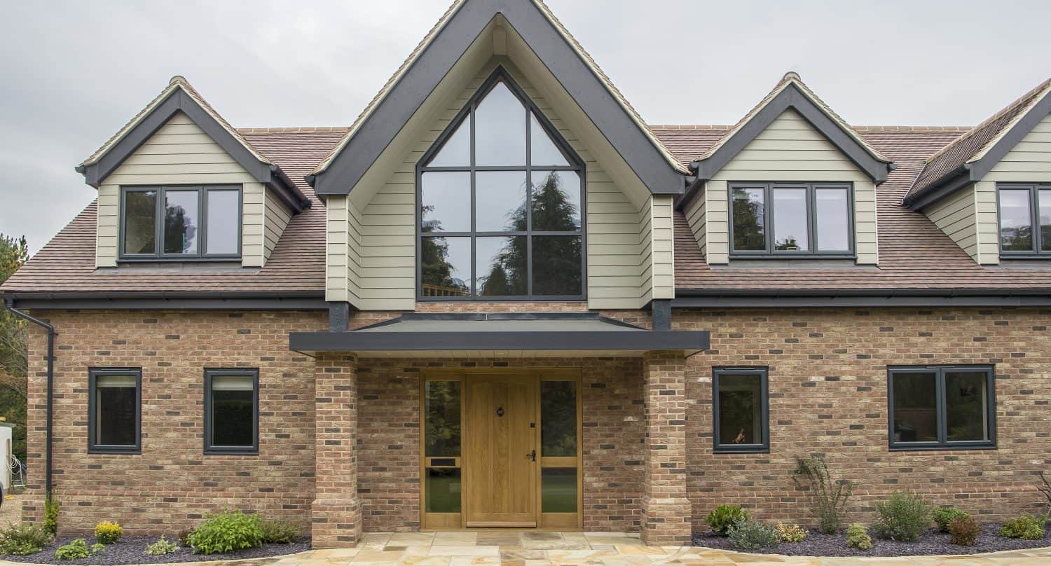 Modern two-story house with brick exterior, large triangular window above the wooden front doors, black-framed windows, sliding doors, and a landscaped front yard with grass and plants.