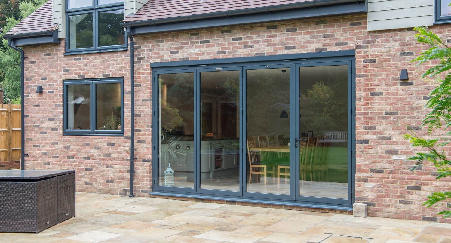 A modern brick house with large glass sliding patio doors and a window, opening onto a stone-tiled patio with outdoor furniture, and reflections of trees visible in the glass.