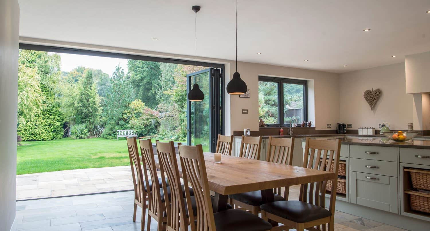 A modern dining area with a wooden table and chairs, pendant lights, and large glass doors open to a lush green garden, allowing natural light to fill the bright, airy space.