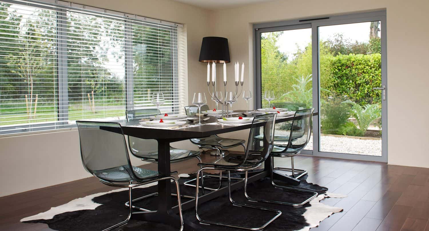 A modern dining room with a black table set for six, transparent chairs, and a black cowhide rug. Large windows and a glass door offer views of a green, leafy garden outside.