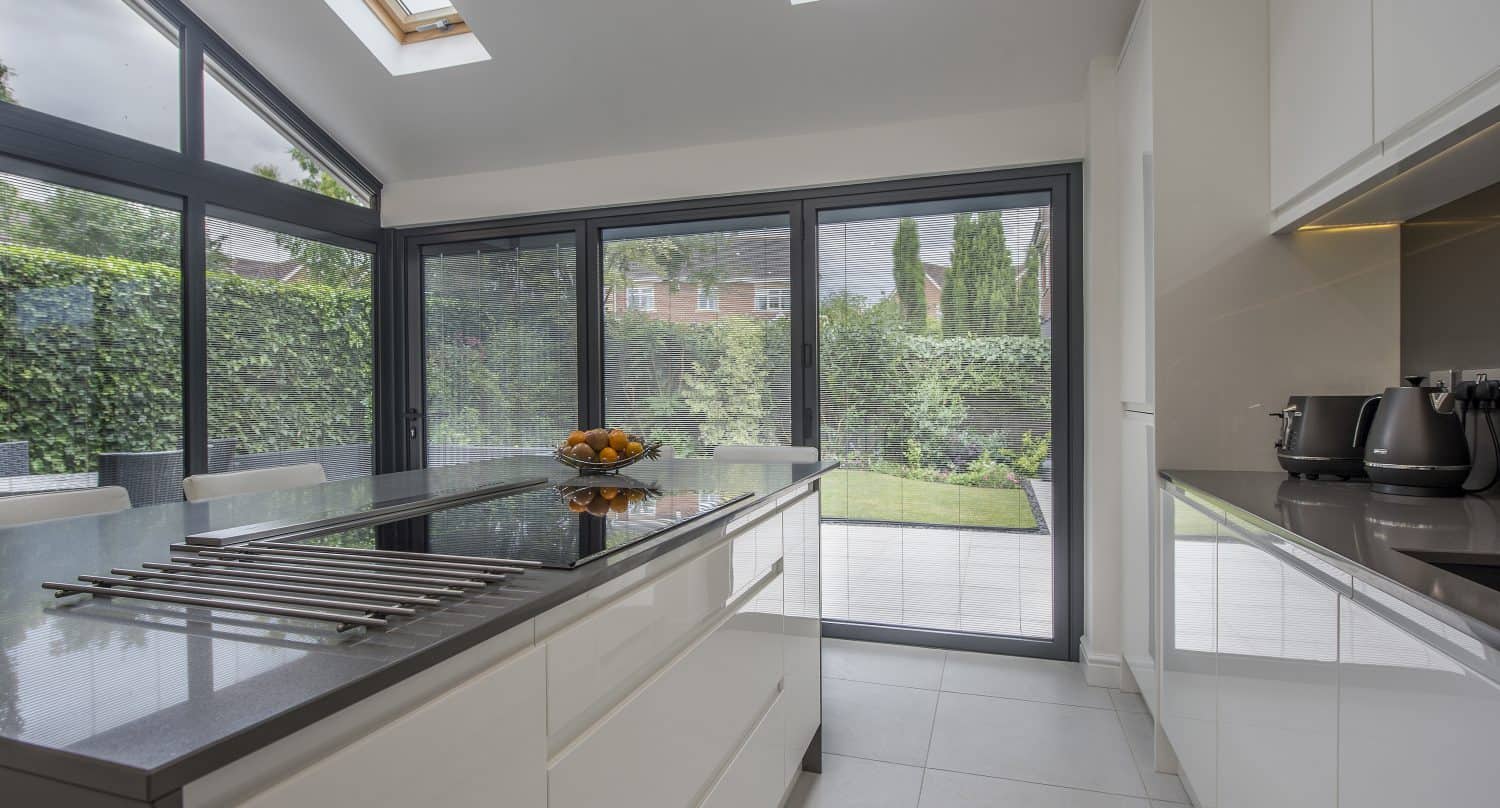 Modern kitchen with white cabinets, an island with a built-in stove, and large bifold doors opening to a green garden. Skylights and windows let in natural light, creating a bright, airy space enhanced by bespoke glass solutions.